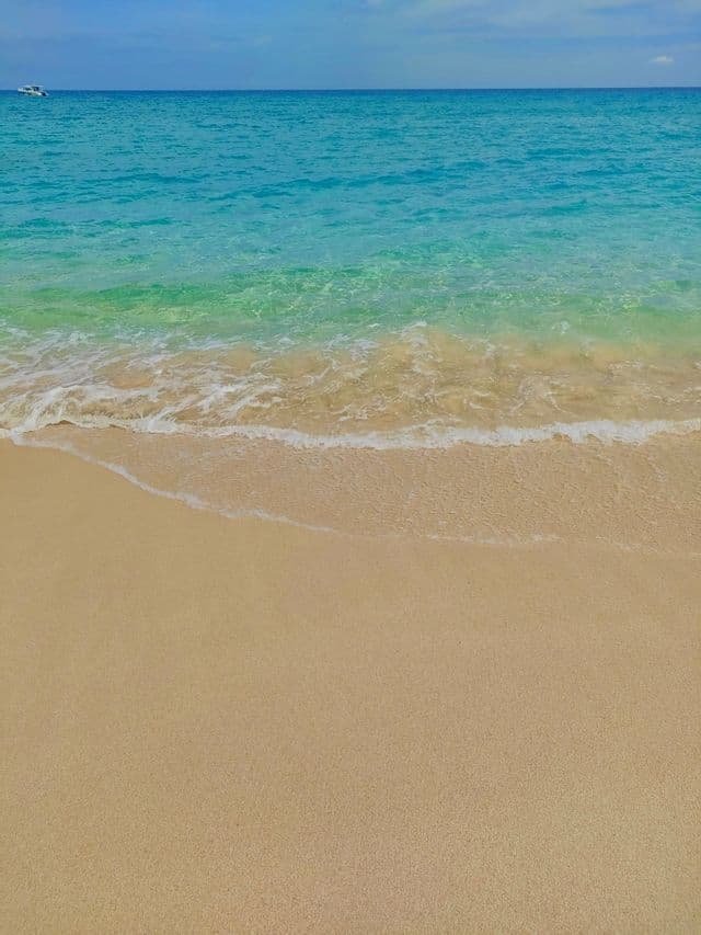 Una suave ola de agua turquesa cristalina baña una playa de arena bajo un cielo azul, con un pequeño bote en el horizonte.