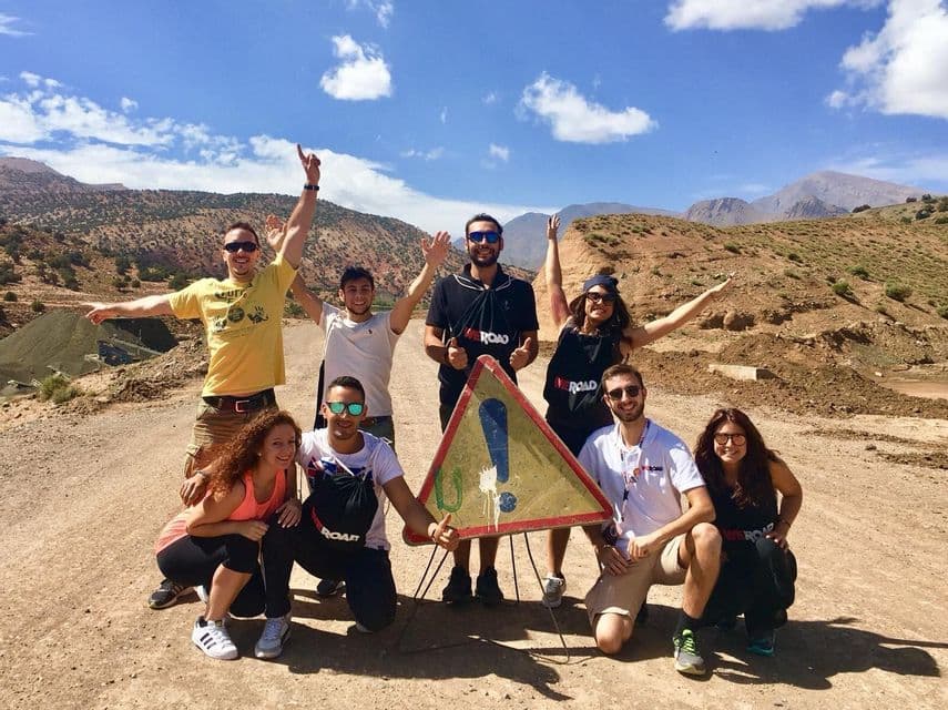 Un viaggio di gruppo WeRoad con otto persone in posa attorno a un segnale stradale triangolare su una strada sterrata, con le montagne sullo sfondo.