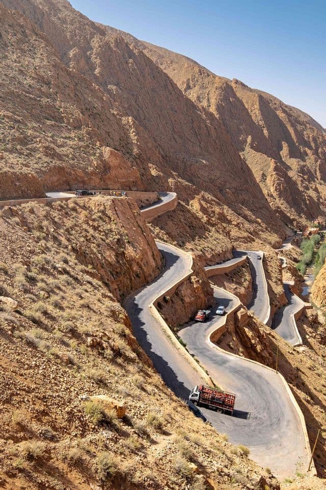 Luftaufnahme einer kurvenreichen Straße mit Serpentinen, die sich eine steile, felsige Bergschlucht unter klarem Himmel hinabwindet, mit Autos und einem Lastwagen darauf.