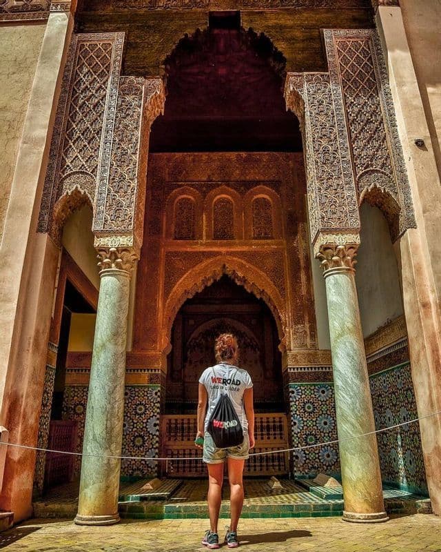 A woman wearing a WeRoad t-shirt and backpack stands looking up at a large, ornate archway with detailed carvings and tile mosaics.