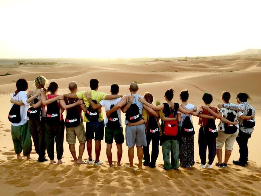 A WeRoad group trip standing arm-in-arm on a sand dune, looking out over a vast desert landscape at sunset.