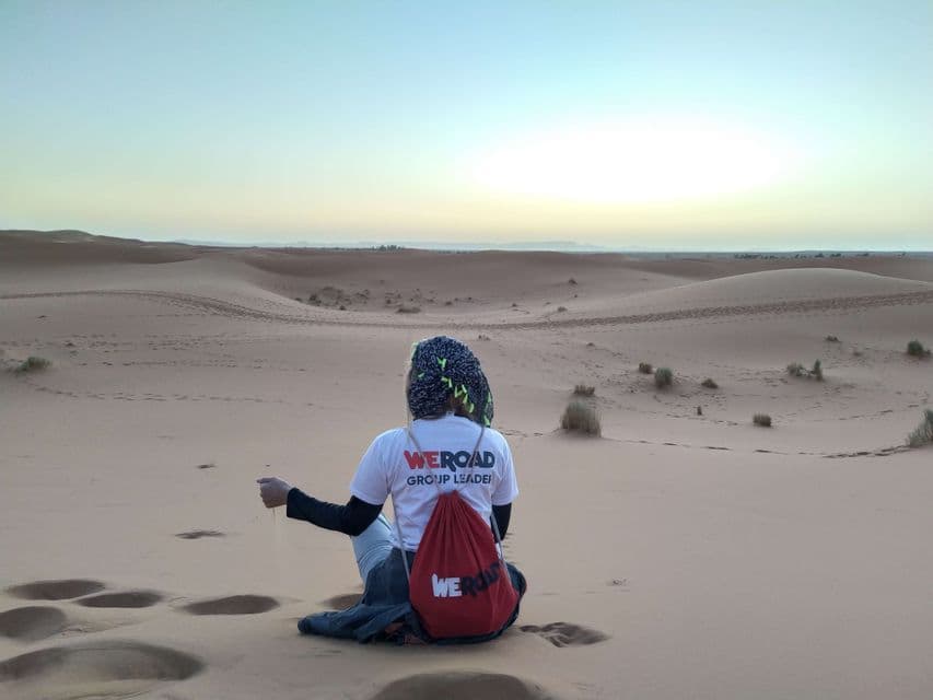 A WeRoad group leader sits on a sand dune, letting sand fall from their hand while looking at the desert landscape at sunset.