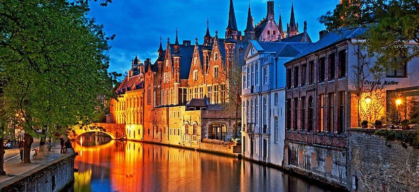 Des bâtiments historiques en briques avec des flèches sont illuminés le long d'un canal au crépuscule, leurs lumières se reflétant dans l'eau.