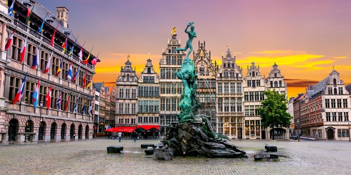 Une fontaine en bronze ornée sur une place pavée entourée de bâtiments historiques décorés de drapeaux sous un ciel de coucher de soleil.