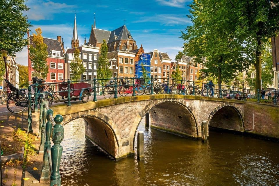 Des vélos sont garés sur un pont de briques en arc au-dessus d'un canal, avec des bâtiments européens historiques et des arbres sous un ciel bleu.