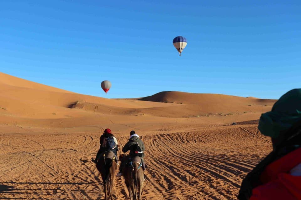 A WeRoad group trip rides camels across desert sand dunes as two hot air balloons float in the blue sky.