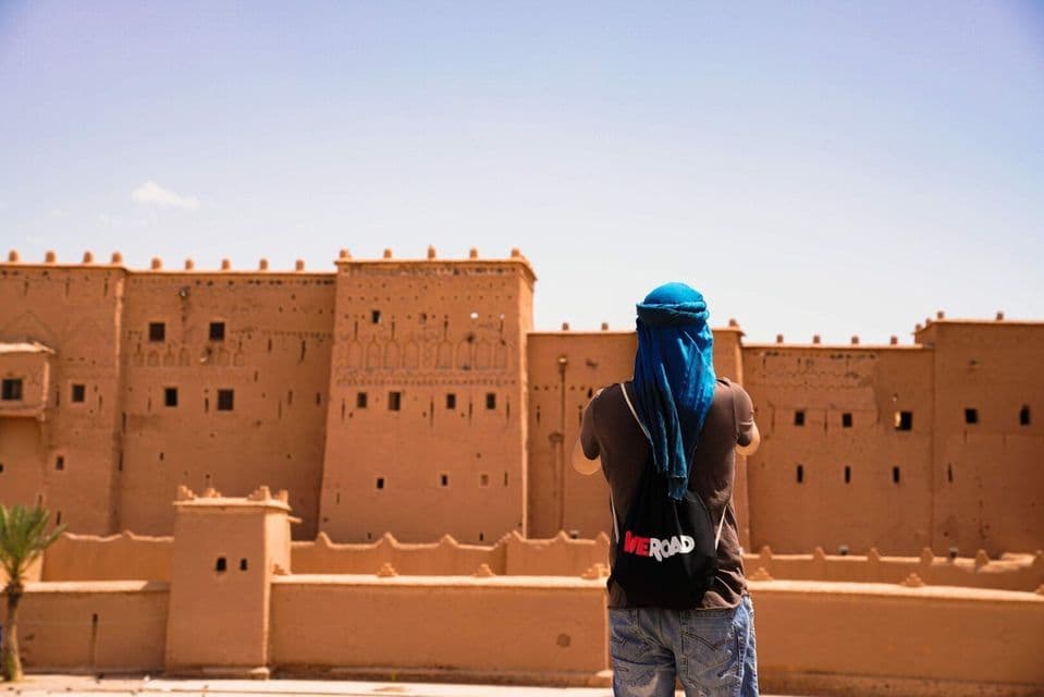 A person from behind, wearing a blue headscarf and a WeRoad backpack, faces a large, sand-colored kasbah under a clear sky.
