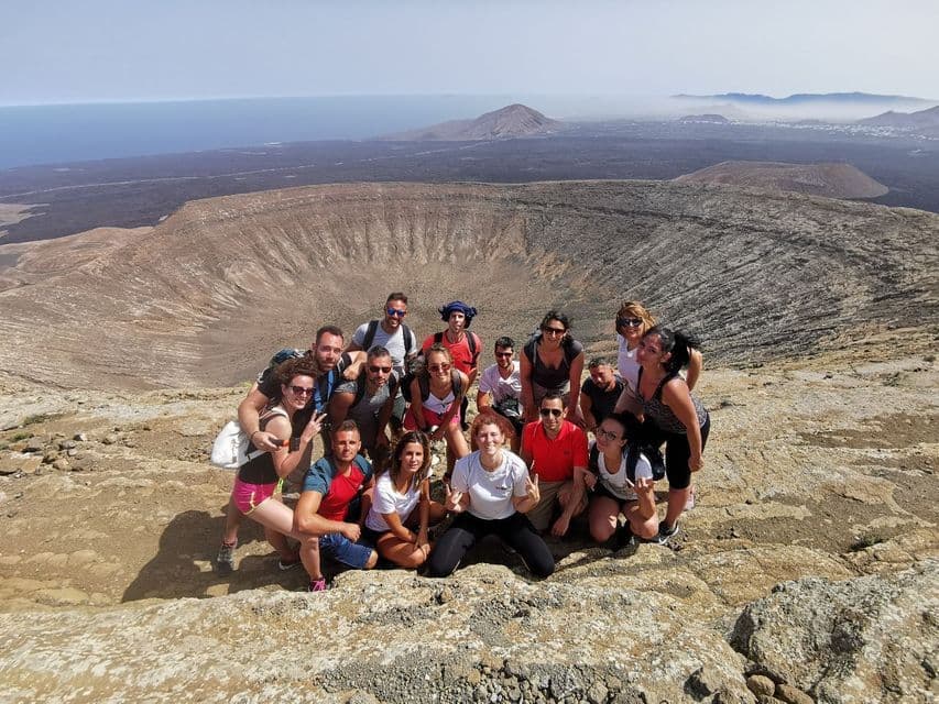 Eine WeRoad-Gruppe posiert für ein Foto am felsigen Rand eines großen Vulkankraters, im Hintergrund Lavafelder und der Ozean.