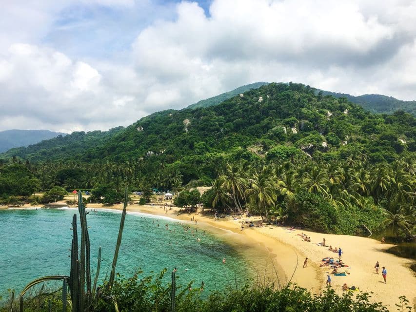 Ein erhöhter Blick auf einen tropischen Strand, wo Menschen im türkisfarbenen Wasser schwimmen, mit einem großen, bewaldeten Hügel im Hintergrund.