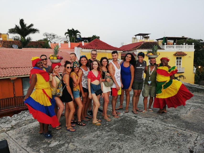 Un viaje en grupo de WeRoad posa para una foto en una azotea con dos mujeres vistiendo trajes tradicionales coloridos y equilibrando fruta en sus cabezas.
