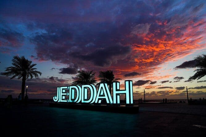 Une grande enseigne lumineuse affichant 'JEDDAH' sur une promenade, avec des palmiers en silhouette devant un ciel de coucher de soleil coloré.