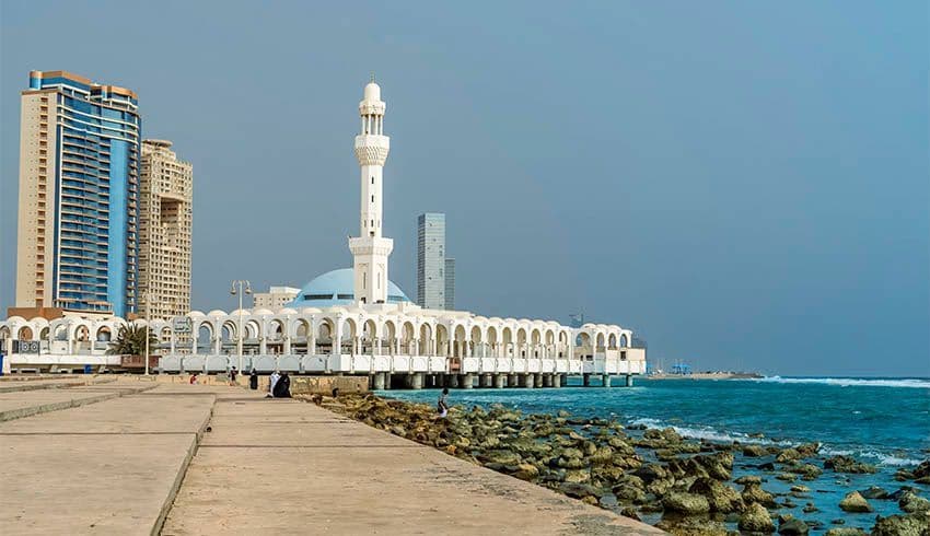 Une mosquée blanche avec un dôme bleu et un minaret est située sur une jetée au-dessus de la mer, avec un littoral rocheux et des gratte-ciel modernes à proximité.