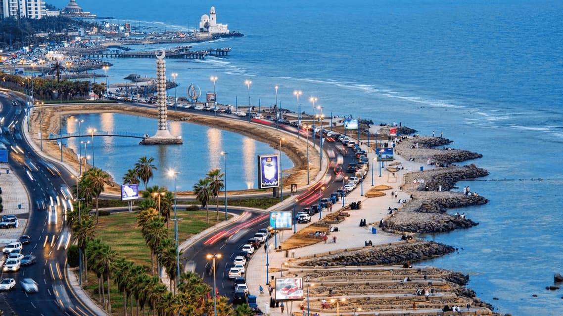 Vue aérienne d'une ville côtière au crépuscule, montrant une autoroute incurvée longeant la mer, avec un grand monument dans une lagune.