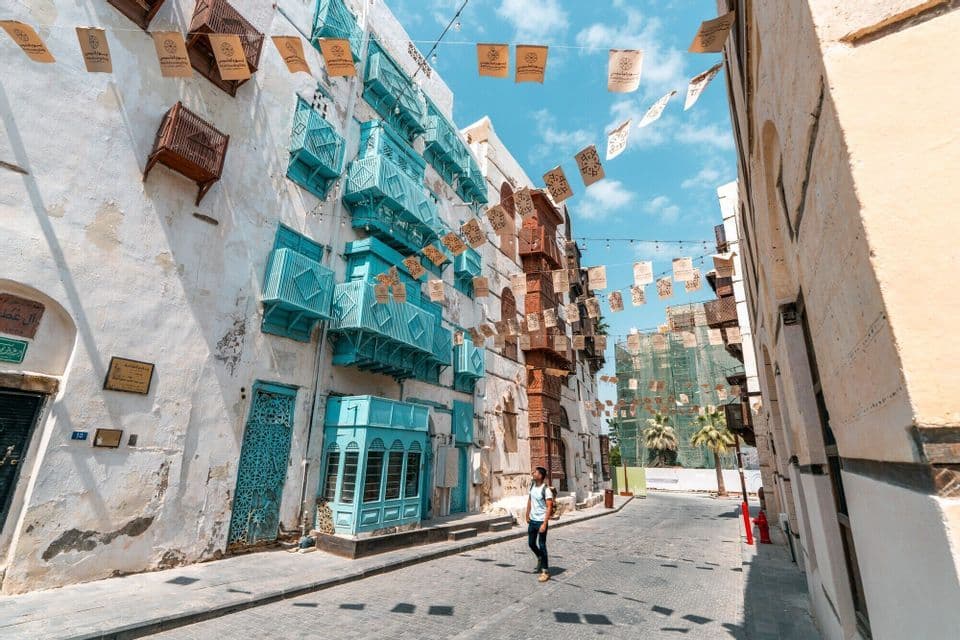 Un homme se tient dans une rue pavée, regardant des bâtiments historiques avec des balcons turquoise vifs et des drapeaux au-dessus.