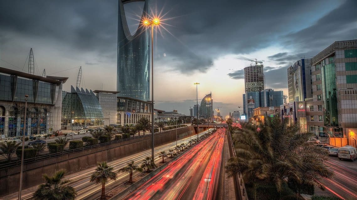 Une autoroute à plusieurs voies, avec des traînées de feux rouges de la circulation, sur fond de gratte-ciel modernes et de palmiers sous un ciel crépusculaire nuageux.