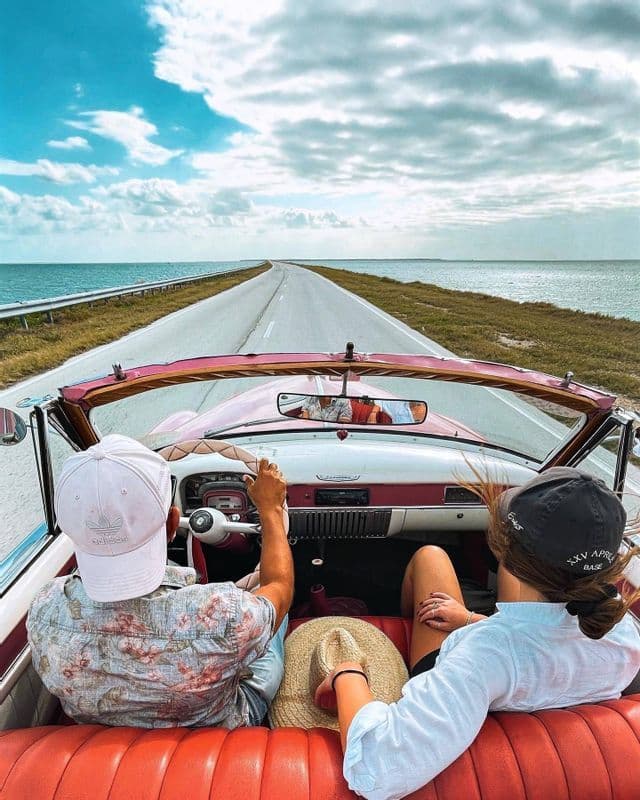 Zwei Teilnehmer einer WeRoad-Gruppenreise fahren in einem pinken Vintage-Cabrio auf einer Küstenstraße am Meer.