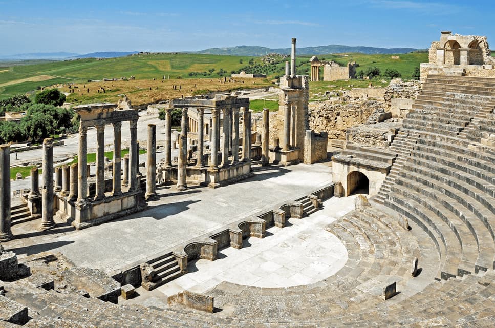 A high-angle view of a large, ancient stone amphitheater and ruins set against a backdrop of green, rolling hills under a blue sky.