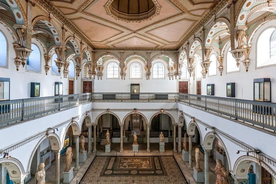 An ornate two-level museum hall viewed from above, with decorative arches, classical statues, and a large mosaic floor.