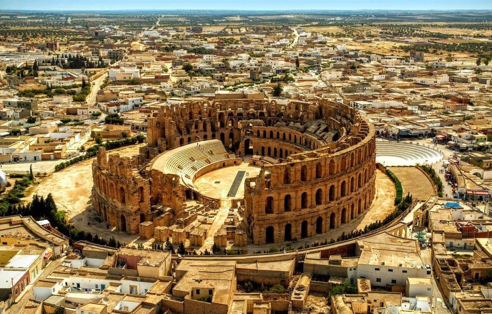 An aerial view of a large, stone Roman amphitheater ruin surrounded by a sprawling town under a clear sky.