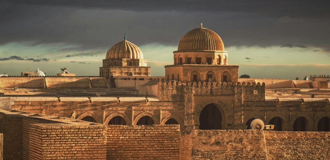 Two domed stone buildings and rooftops of an ancient city are illuminated by sunlight under a dark, cloudy sky.