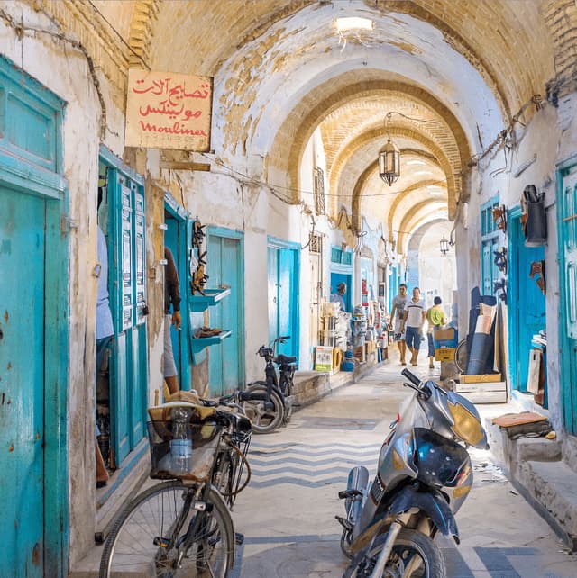 A narrow, covered market alley with stone arches and turquoise doors, with a bicycle and scooter parked in the foreground.
