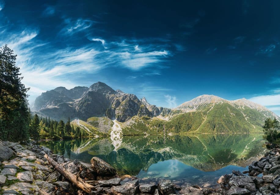 A clear alpine lake perfectly reflects a rocky mountain range under a deep blue sky with wispy clouds, with a stone shore in the foreground.
