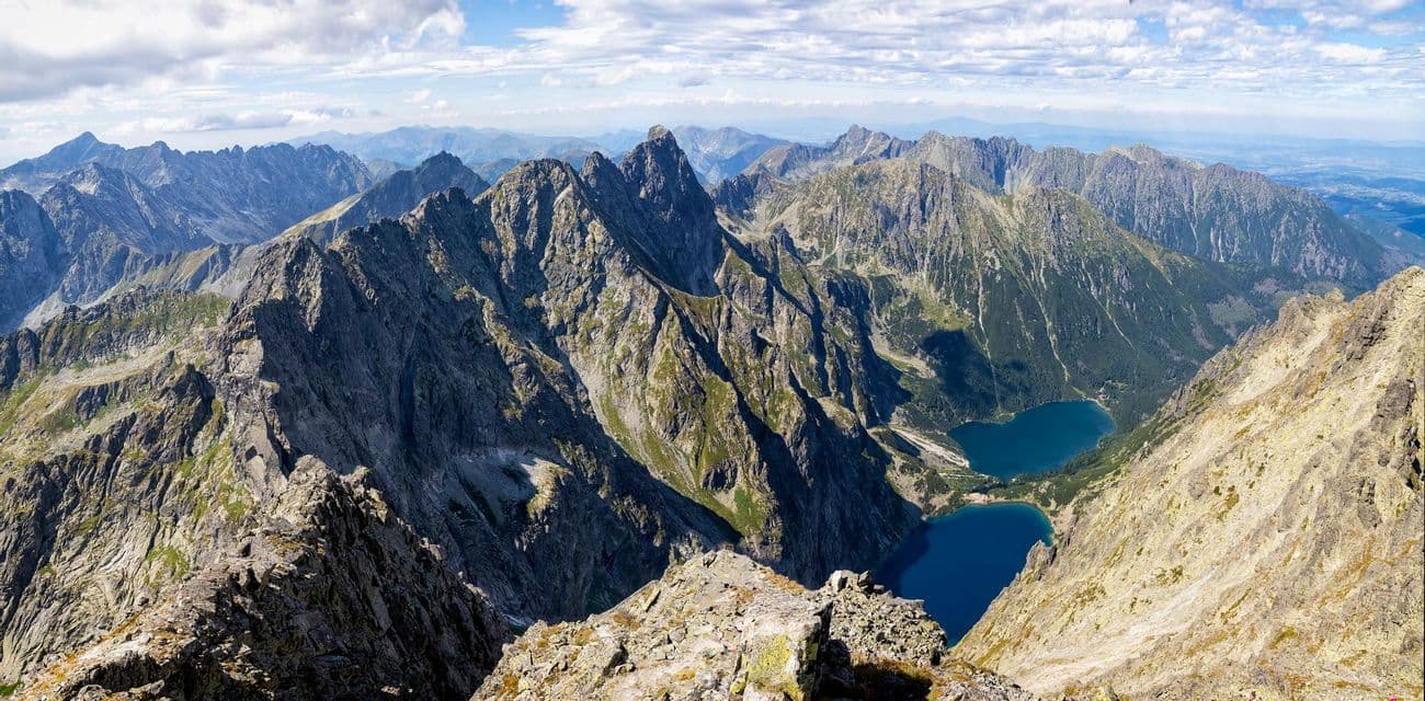 A panoramic view from a rocky summit overlooking a vast mountain range with two blue alpine lakes in the valley.