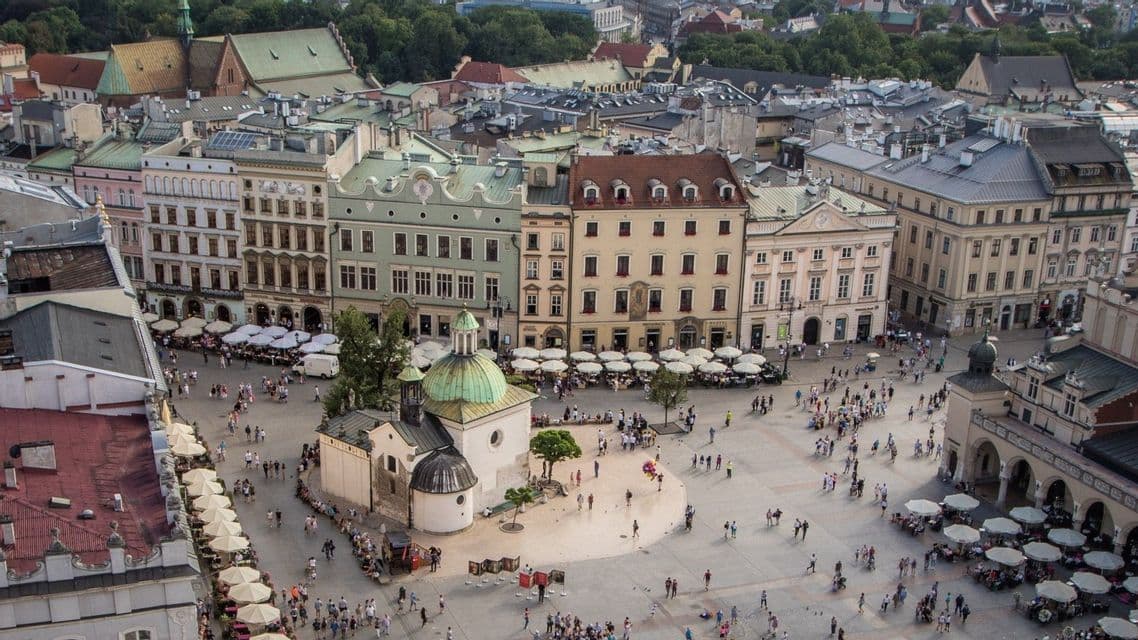 An aerial view of a bustling European city square filled with people, outdoor cafes, and a small church with a green dome.