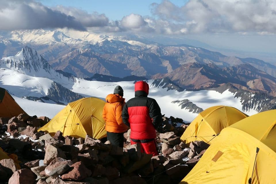 Two people in winter jackets stand at a high-altitude campsite with yellow tents, overlooking a vast panorama of snow-covered mountains.