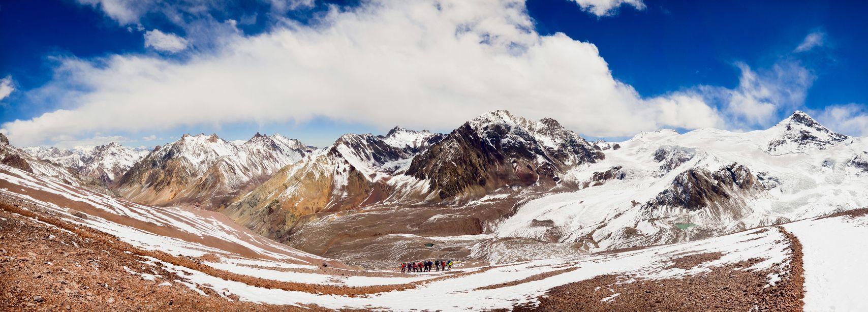 A panoramic view of a WeRoad group trip hiking on a trail in a vast, snow-covered mountain range.