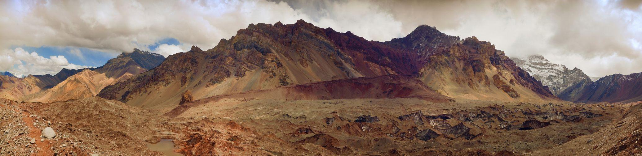 A panoramic view of a rugged, multi-colored mountain range towering over a rocky valley with a debris-covered glacier.