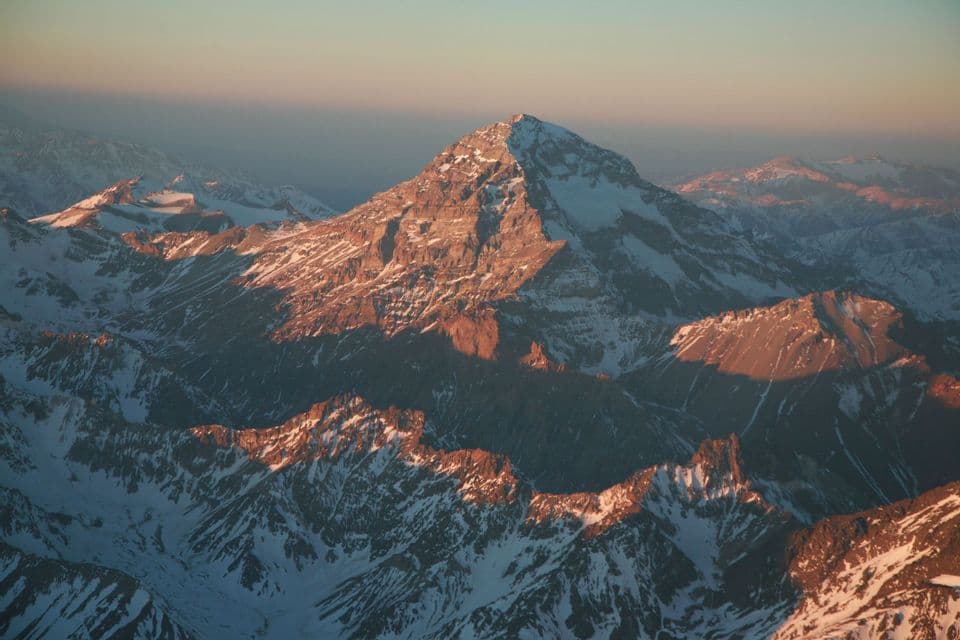 An aerial view of a snow-covered mountain range, with the peaks illuminated by the warm light of a setting sun.