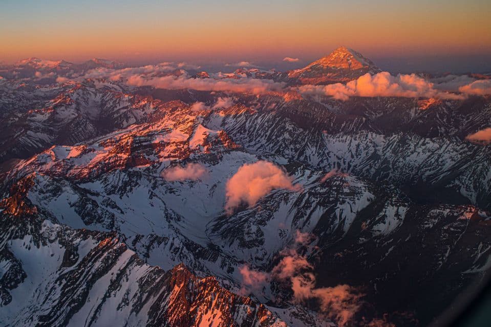 An aerial view of snow-covered mountain peaks and clouds illuminated by the golden light of sunset.