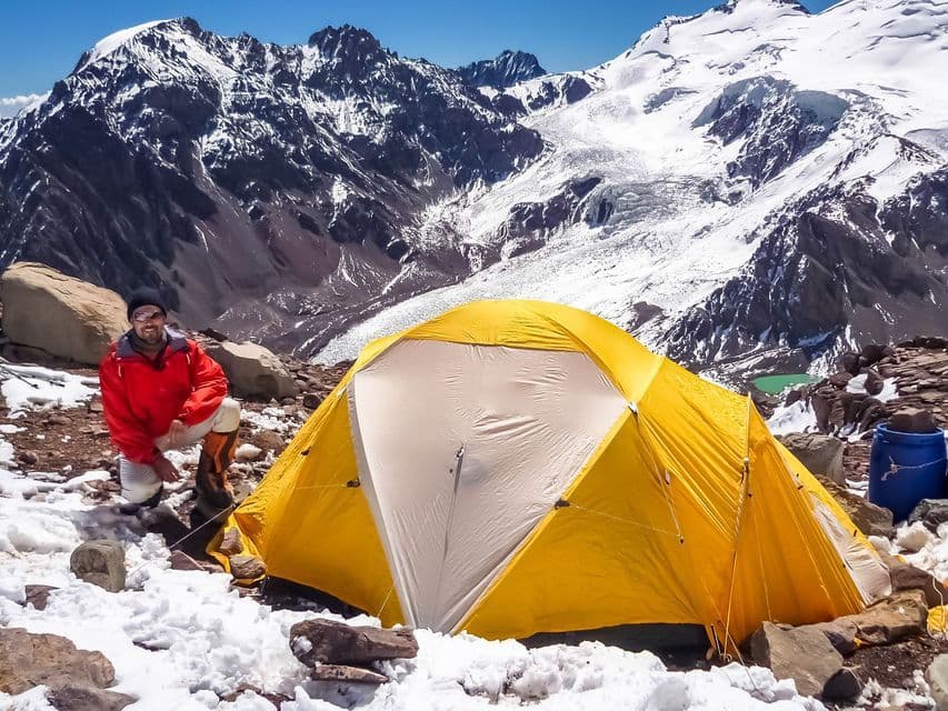 A man in a red jacket crouches next to a yellow tent on a snowy mountain, with a large glacier and jagged peaks in the background.
