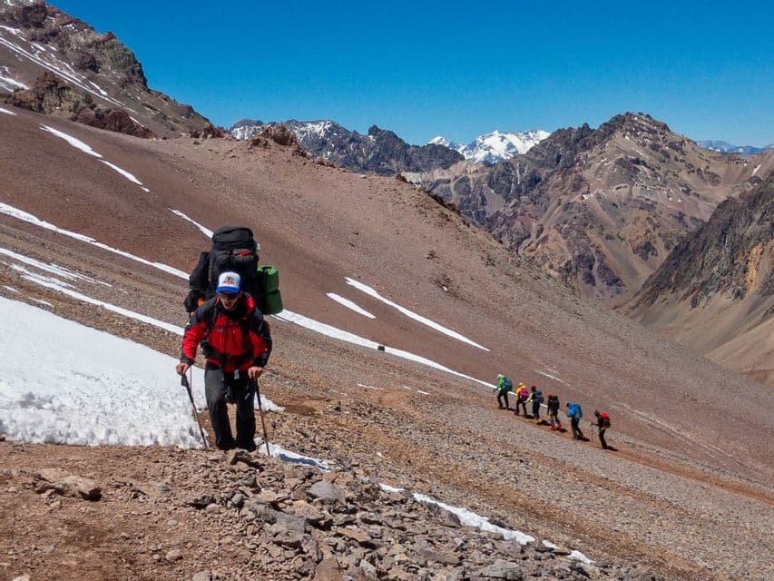 A WeRoad group trip trekking up a steep, rocky mountainside with patches of snow under a clear blue sky.