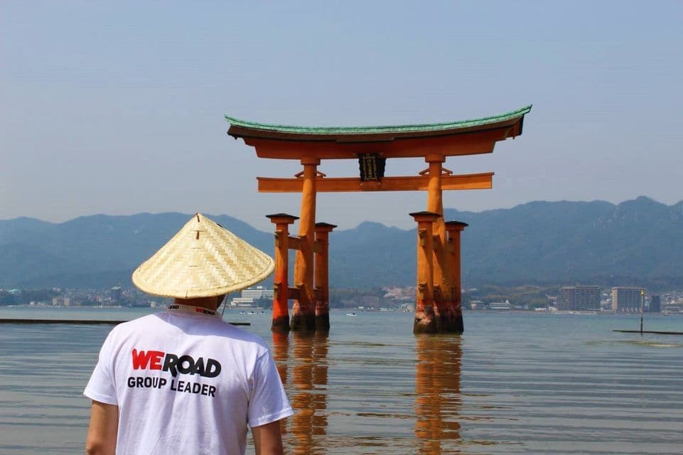 Un capo gruppo WeRoad che indossa un cappello conico osserva un grande Torii arancione eretto nell'acqua, con montagne sullo sfondo.