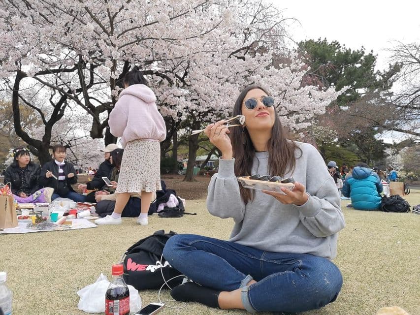 A woman in sunglasses eats sushi with chopsticks while sitting on the grass for a picnic under a blooming cherry blossom tree.