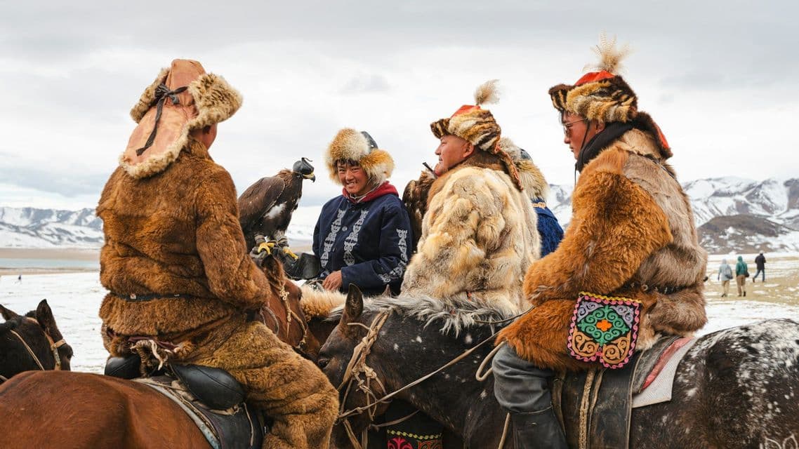 A group of eagle hunters in traditional fur clothing sits on horseback, with an eagle, against a backdrop of snowy mountains.