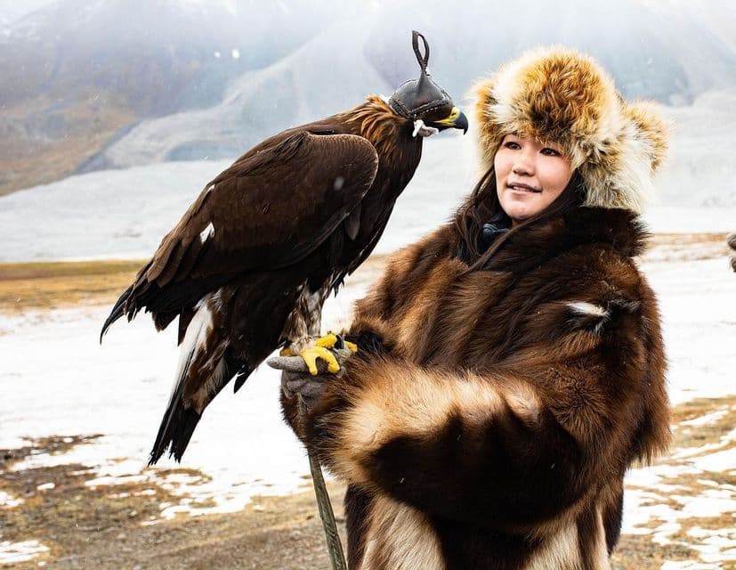A person in a fur coat and hat holds a hooded eagle on their gloved hand in a snowy mountain landscape.