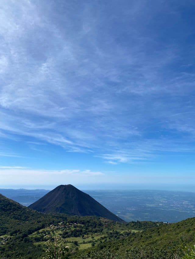 Ein dunkler Vulkankegel erhebt sich hoch inmitten saftig grüner Hügel und überblickt eine weite Ebene unter blauem Himmel mit leichten Wolken.