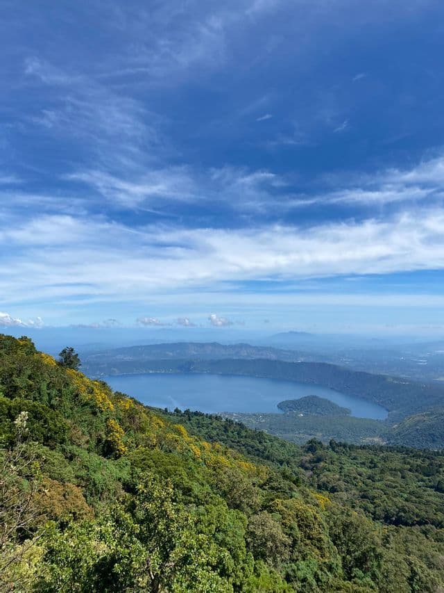 Eine Vogelperspektive auf einen großen Kratersee, umgeben von bewaldeten Bergen unter einem weiten blauen Himmel mit zarten Wolken.