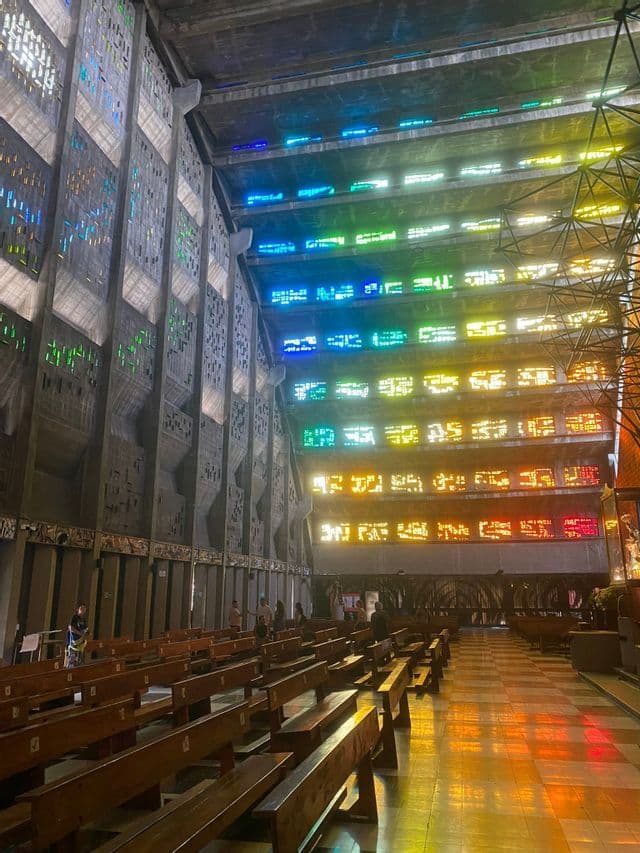 Buntes Licht von Buntglasfenstern beleuchtet die Kirchenbänke und den karierten Boden in einer riesigen, modernen Betonkirche.
