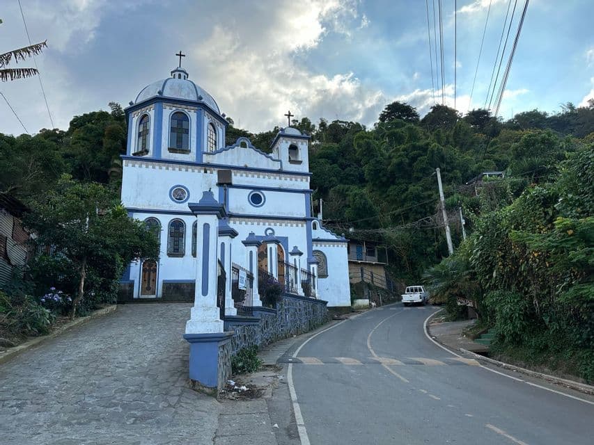 Eine weiß-blaue Kirche mit einer Kuppel steht auf einem Kopfsteinpflasterweg neben einer gewundenen Straße an einem bewaldeten Hang.
