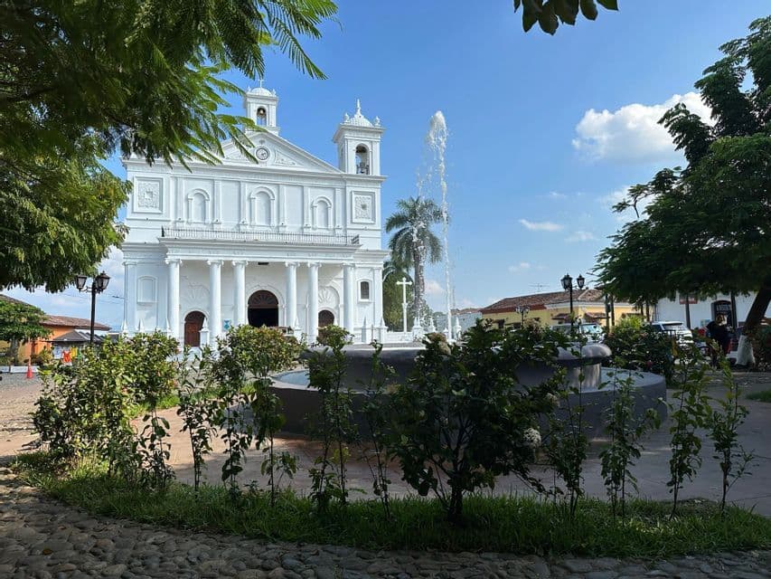 Eine große weiße Kirche mit zwei Glockentürmen und klassischen Säulen steht hinter einem öffentlichen Platz mit einem Brunnen und grünen Büschen.