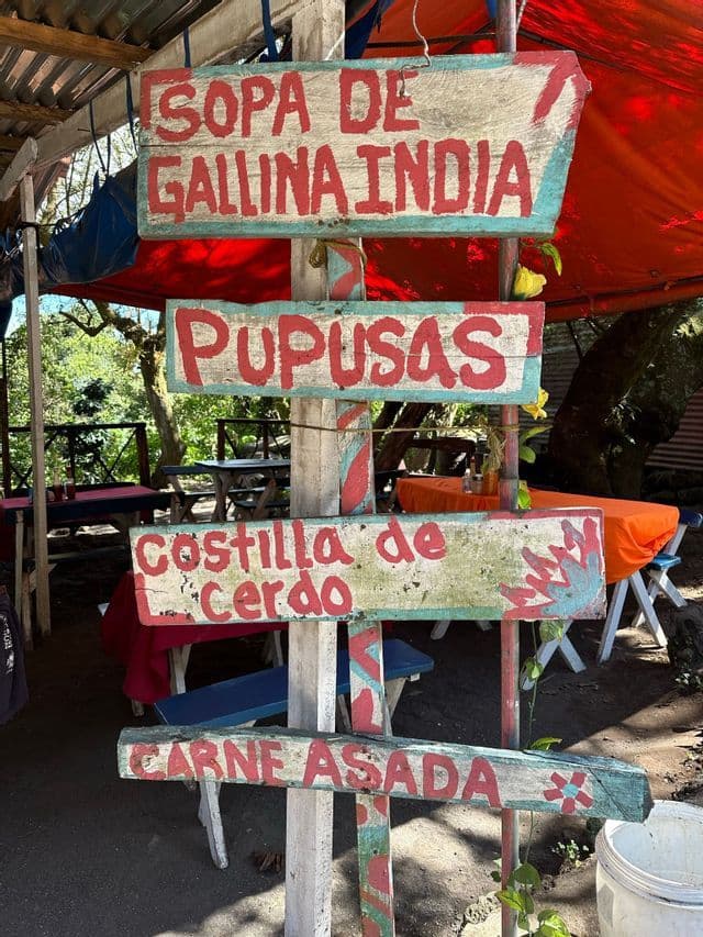 Handbemalte Holzschilder an einem Pfahl bewerben ein spanisches Menü mit Pupusas und Carne Asada an einem rustikalen Imbissstand.