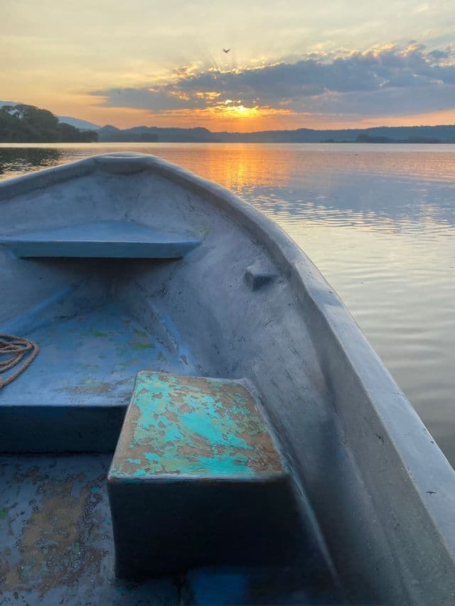 Blick vom Bug eines kleinen Bootes auf einem ruhigen See, während die Sonne über fernen Hügeln aufgeht.