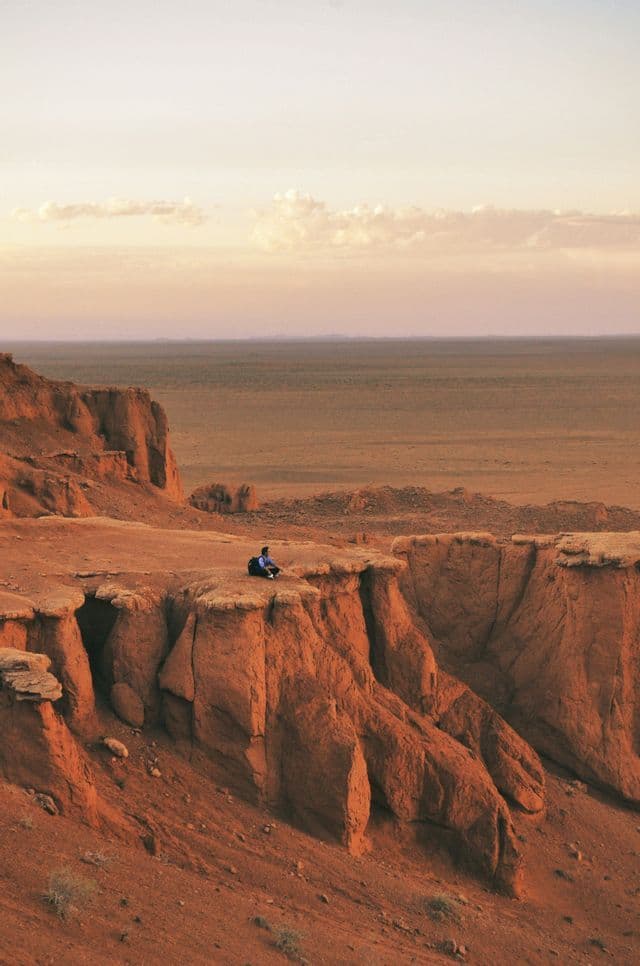 Eine Person mit einem Rucksack sitzt am Rande eines roten Felsencanyons und blickt während eines blassen Sonnenuntergangs über eine weite Wüstenebene.