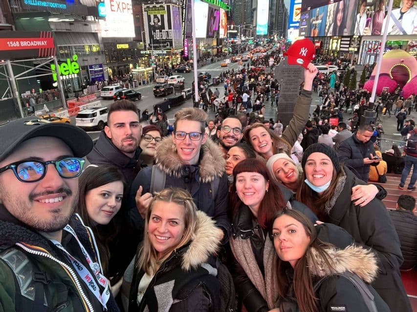 A WeRoad group trip takes a selfie on steps overlooking a bustling city square filled with bright billboards and crowds.