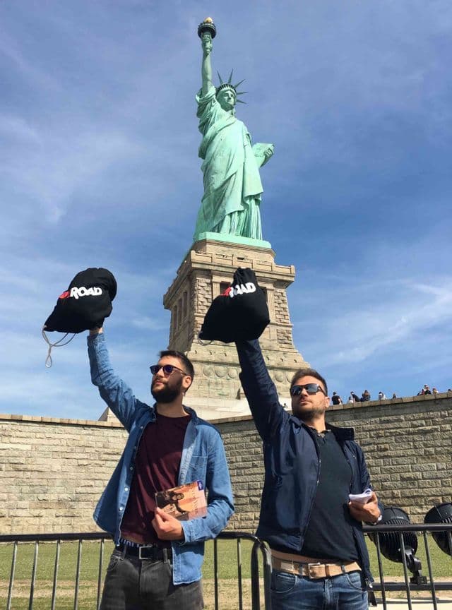 Dos hombres de un viaje en grupo de WeRoad sosteniendo bolsas negras frente a la Estatua de la Libertad bajo un cielo azul.