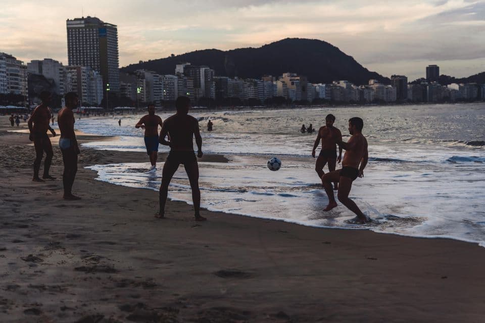 Un grupo de hombres silueteados jugando al fútbol en la orilla de una playa al atardecer, con el horizonte de una ciudad de fondo.